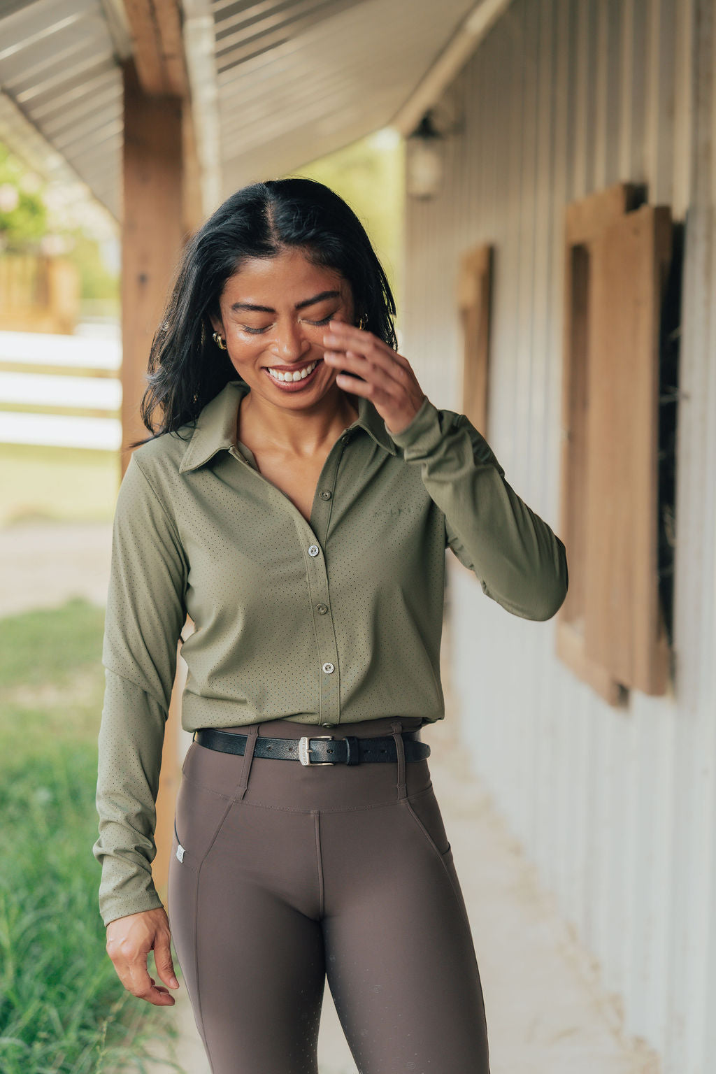 A woman smiling on a porch wearing an olive green long-sleeve button-up shirt and brown riding breeches.