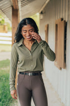 A woman smiling on a porch wearing an olive green long-sleeve button-up shirt and brown riding breeches.