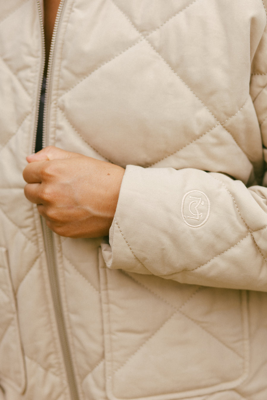 Close-up detail shot of a cream-colored quilted jacket sleeve featuring diamond-pattern stitching and a circular embroidered logo patch, with a front zipper visible in the backgroun