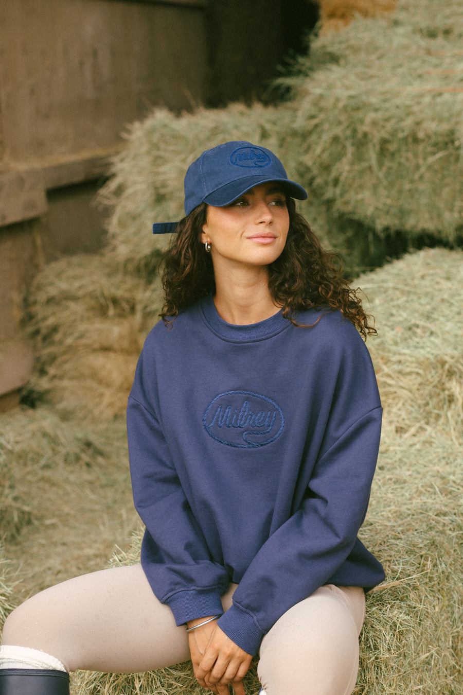 Woman with curly brown hair wearing a navy blue crewneck sweatshirt with embroidered logo and matching navy baseball cap with logo embroidery, standing outdoors against a hay bale background
